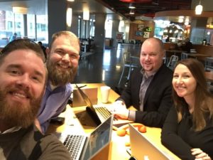 A group photo image of Director of Engineering, Greg Rickaby, Lead Backend Engineer, Ben Lobaugh, CEO, Brad Williams, and Director of Project Management, Cristina Holt, smiling at the camera while sitting in a coffee shop with their laptops open.