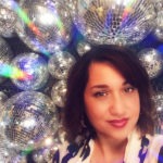 A photograph of WebDevStudios Communications Specialist, Laura Coronado, looking into the camera smiling wihile standing in front of wall of disco balls.