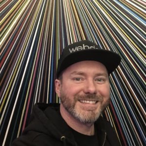 A photograph of WebDevStudios engineer, Jeremy Ward, smiling and wearing a WebDevStudios baseball cap while standing in front of a wall painted with various colored stripes at Meow Wolf in Santa Fe, New Mexico.
