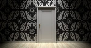 Photograph of a wall with black and white decorative wallpaper and a white door, which is shut, in the center of the wall.
