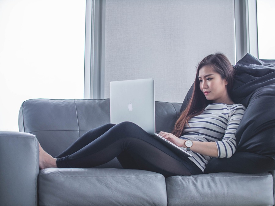 Photographic image of a woman laying on a gray sofa with her Mac Book in her lap as she looks at something unknown on the monitor used as an image for the blog post, "Web Design and Development Myths 2019 Edition."