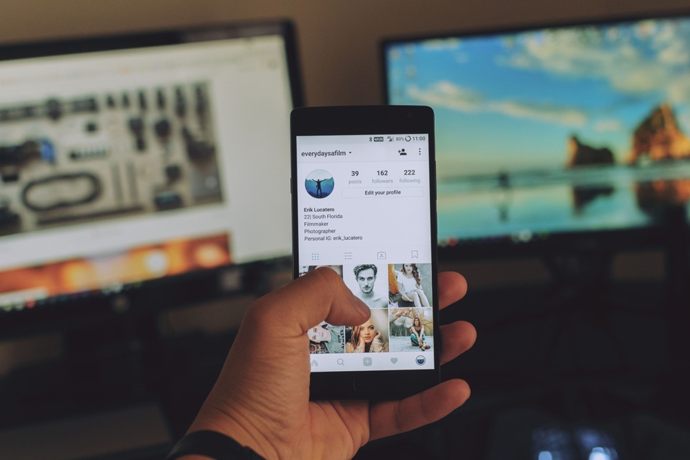 Photo image of a person holding a smartphone with the social media platform Instagram open while there are two computer monitor screens blurred in the background.