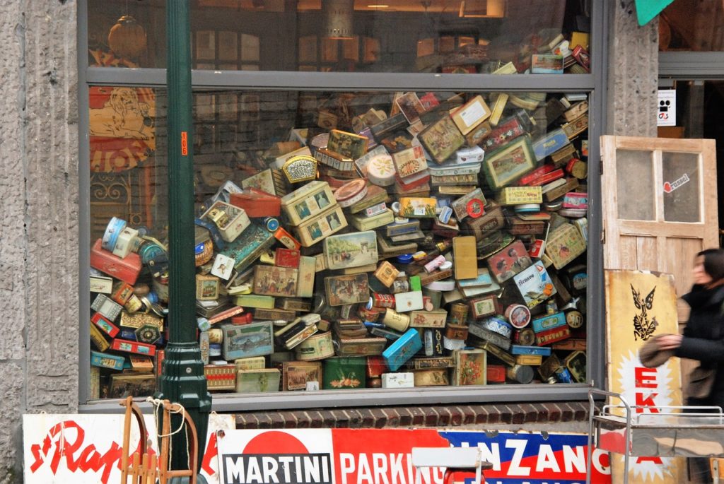 One of the rules of redesign is that there is such a thing as too much. This image is a photograph of a storefront window of a junk shop and shows an extreme amount of vintage tins stacked atop of one another, with the pile falling over.