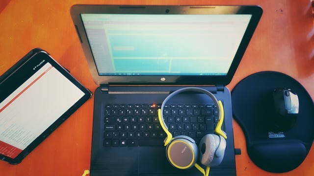 A photographic still shot of an opened laptop with the WordPress dashboard on the screen, a pair of headphones sitting on the keyboard of the laptop, a tablet sitting to the left, and a pad and mouse sitting to the right. All of the items are atop a brown table.
