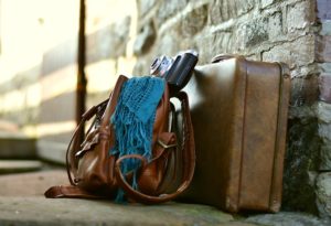 A photograph of a small suitcase sitting on a sidewalk and leaning against a brick wall with a brown leather satchel leaning against it and a blue scarf and camera sitting on top of the satchel.