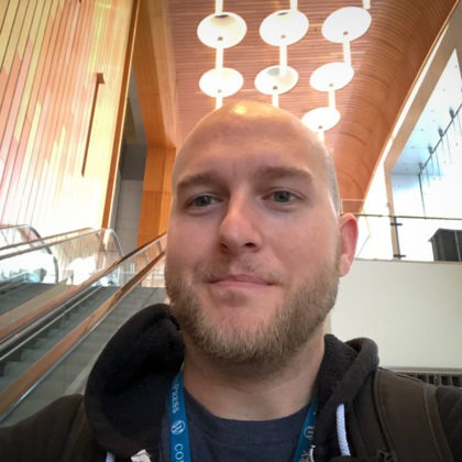 A photo head shot of Mike England, Frontend Engineer at WebDevStudios. he's slightly smiling at the camera while riding down an escalator and wearing a WordPress WordCamp lanyard.