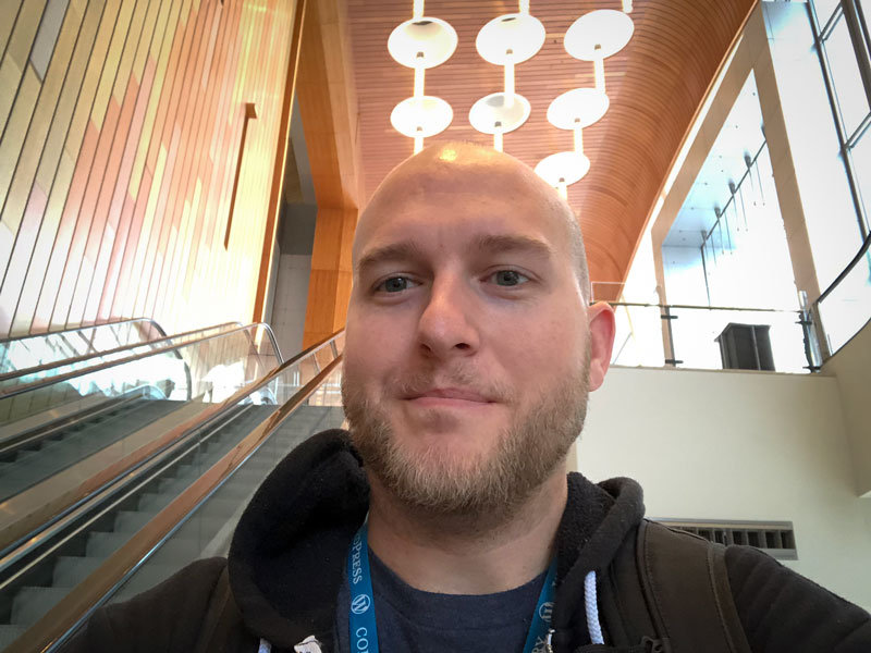 A photo head shot of Mike England, Frontend Engineer at WebDevStudios. he's slightly smiling at the camera while riding down an escalator and wearing a WordPress WordCamp lanyard.