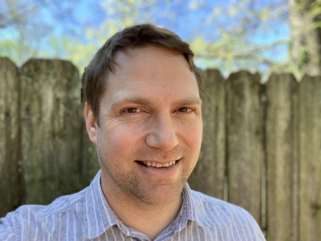A photograph head shot of Marty O'Connor, a frontend engineer at WebDevStudios. He is smiling at the camera and is posed outdoors in front of a wood fence with a blue sky behind.