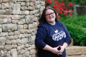 A portrait photograph of Technical Project Manager, Meagan Hanes, leaning against a cobblestone wall and smiling at the camera. Meagan is speaking at WordCamp Hamilton on June 1st.