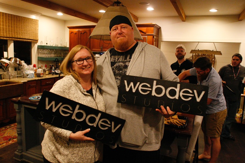 A photograph of COO, Lisa Sabin-Wilson, and Lead Frontend Engineer, Will Schmierer, standing side-by-side and smiling at the camera as the both hold large WebDevStudios plaques, which Will handcrafted himself from wood.