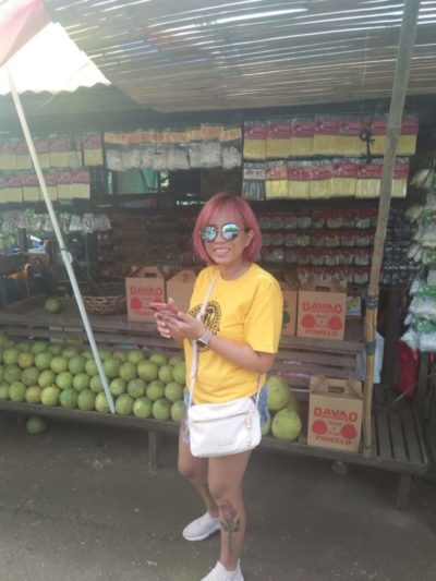 A photograph of WebDevStudios Frontend Engineer, JC Palmes, as she poses and smiles at the camera while standing in front of a fruit stand. She is wearing a yellow T-shirt, jean shorts, sunglasses, and a white crossbody bag.