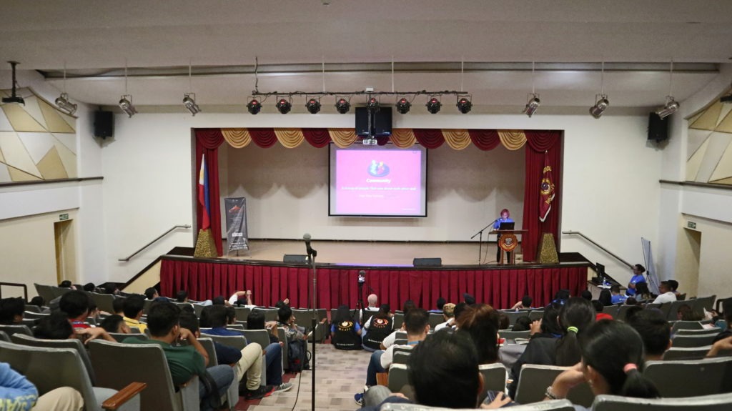 A photograph taken from the top level of an auditorium-style classroom with attendees in the seats listening to WebDevStudios Frontend Engineer, JC Palmes, as she speaks on stage at a podium during WordCamp Davao.