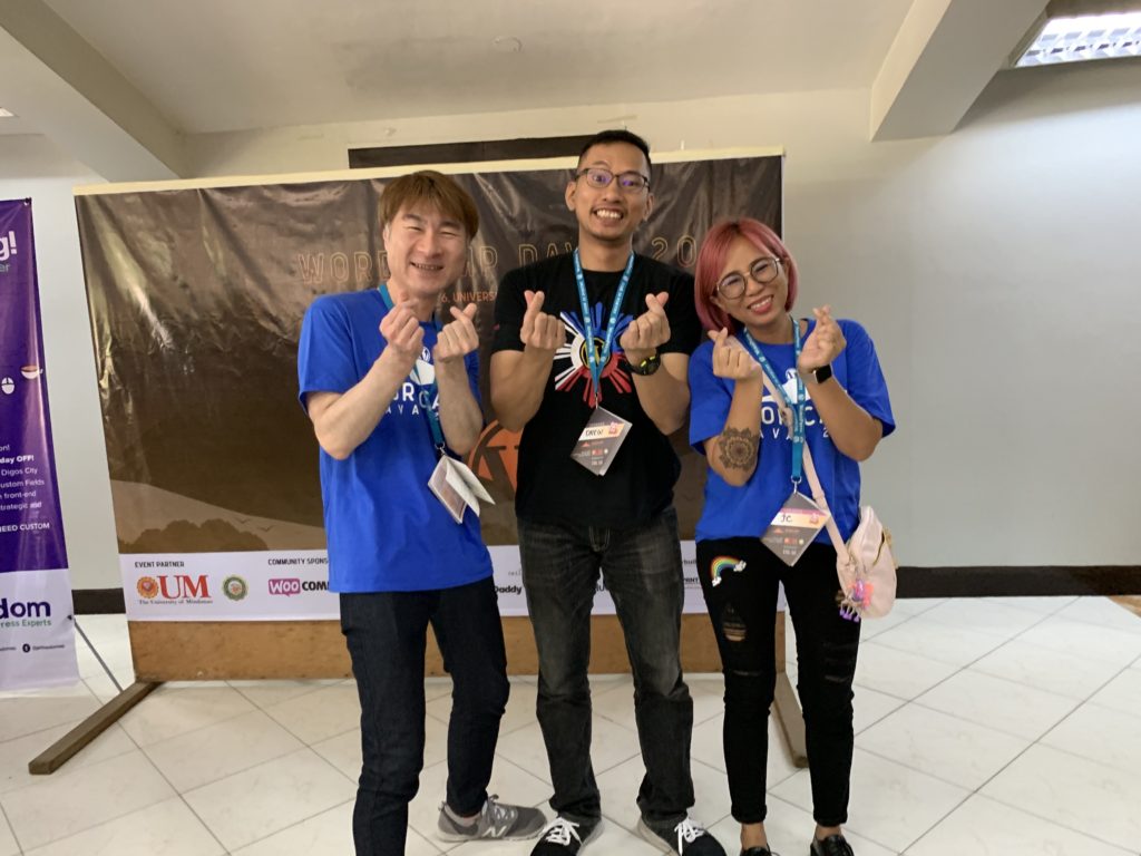 A photograph of WebDevStudios Frontend Engineer, JC Palmes, posing and smiling with Andrew Garcia, lead organizer of WordPress do_action Manila 2019 and Takayuki Miyoshi, creator of Contact Form 7 while they attend WordCamp Davao. They are also holding both hands up as they snap their fingers.