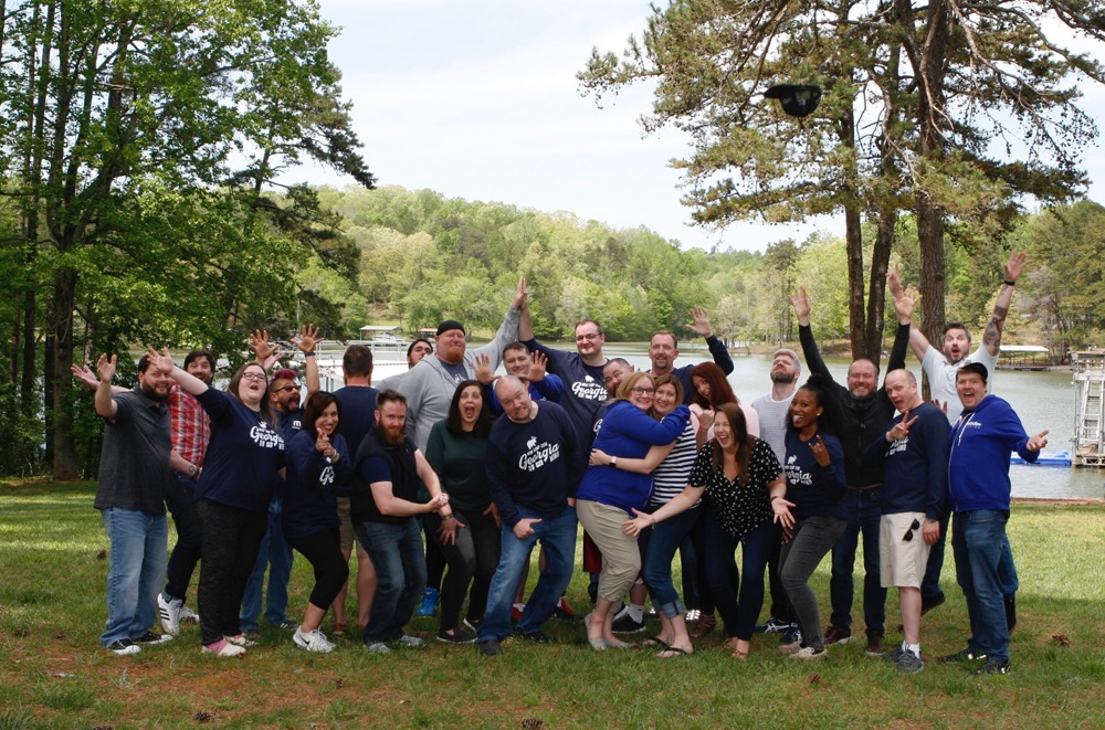 This an outdoor photograph of the WebDevStudios team, taken at WDS Camp 2019 at Lake Lanier in Georgia. Each individual team member is posing in a silly manner nearby the lake.
