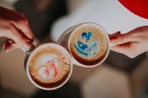 This is an image of a photograph taken from above looking down on two different hands each holding a cup of coffee, both with decorative foam shaped in leaves. This image is aligned left within the blog post's paragraph with the text of the paragraph wrapping around the image.