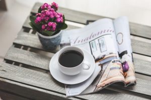 A photo image of a cup of coffee sitting atop a magazine with a pretty purple floral plant in a small pot sitting next to the coffee and magazine.
