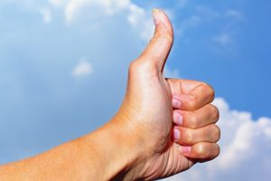 This is a photo of a person's hand giving the thumbs up sign against a blue sky with white clouds in the background.