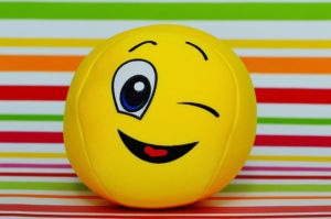 This is a photo of a yellow ball with the winky face emoji printed on it. The ball sits against a multi-color, striped background.