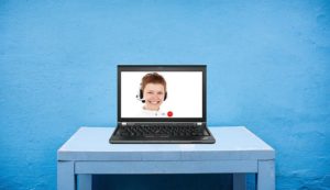 A photo of a bright blue wall with a white desk sitting in front of it and an open laptop sitting on top of the desk displaying a woman smiling while wearing a headset as though she is conducting a live online meeting.