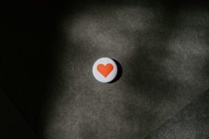 A photograph of a pin-on button of a red heart against a white background and the button is laying on a cement ground.