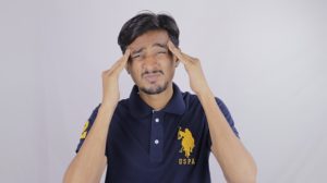 This is a photograph of a man wearing a navy polo style shirt and making a face as though he's in pain. His hands are at his temples as though he has a headache.