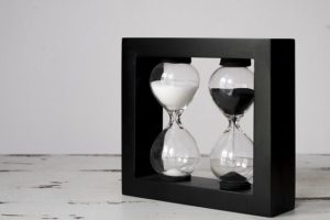 This is a photograph of two hourglasses filled with sand, one white, one black. They are framed by a black frame.