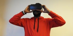 This is a photograph of a man wearing a red hoodie shirt and a virtual reality technology headset. He has his hands placed on the sides of the headset and he seems preoccupied with whatever he is viewing.
