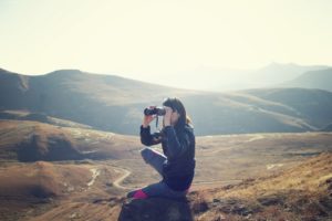 This is a photograph of a woman kneeling and looking through binoculars outdoors at a scenic vieew.