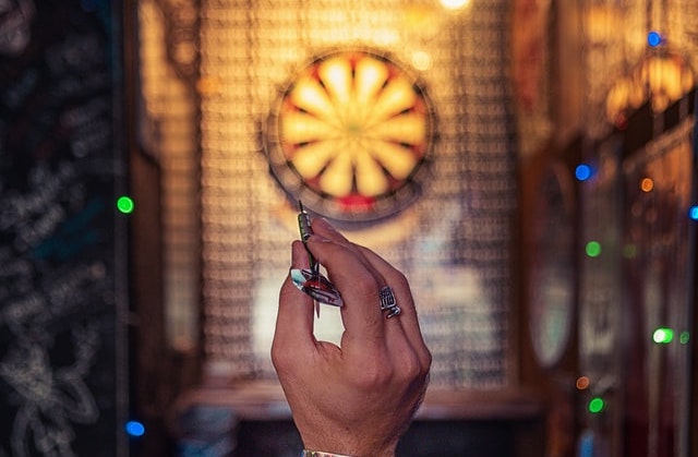This is a photograph of a hand holding a dart and aiming for a dartboard that is blurred in the background.