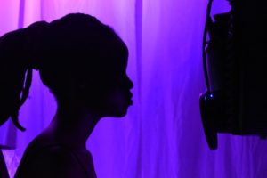 This is a silhouette photograph of a woman at a microphone. Both are backlit by purple light.