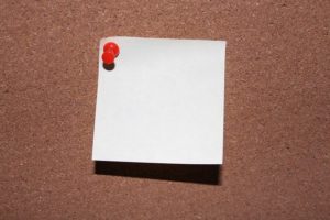 This is a photo of a blank sticky note pinned with a red push pin onto a cork board.