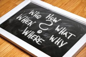 This is a photograph of a small black slate with a white frame and the words, "how, who, what, when, why, where," along with a question mark, written in white chalk on the slate.