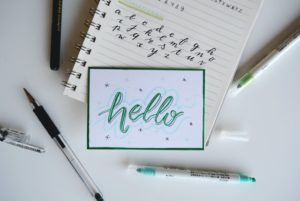 This is a photograph of a white notecard with the word "hello" handwritten on it in green ink.