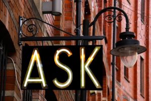 This is a photo of a neon sign hanging on an outdoor wall that says, "Ask."