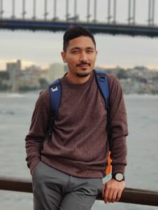 This is a photograph of WebDevStudios Backend Engineer, Alok Shrestha, leaning against a rail, with a water scenery and bridge blurred behind him.