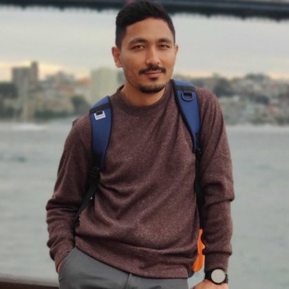 This is a photograph of WebDevStudios Backend Engineer, Alok Shrestha, leaning against a rail, with a water scenery and bridge blurred behind him.