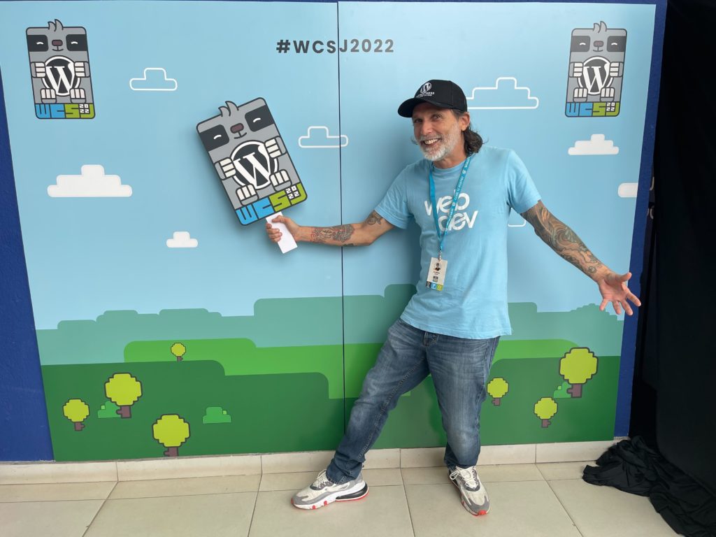This is a photo of Frontend Engineer Alfredo Navas at Word Camp San Jose Costa Rica 2022. He is in front of a step-and-repeat what the WordCamp's logo and he is holding a sign of the WCSJ logo.