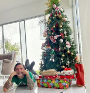 This is a photo of a beautifully decorated holiday tree with Senior Frontend Engineer, Alfredo Navas, laying on the floor next to it, his smiling face propped by his hands.