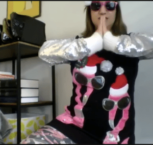 This is a Zoom screenshot of the ugly sweater contest winner, Marketing Manager, Laura Coronado, wearing a black sweater decorated with pink flamingoes wearing Santa hats and sunglasses, and with silver sequin sleeves. She is also wearing pink sunglasses and holding a flamingo yoga pose.