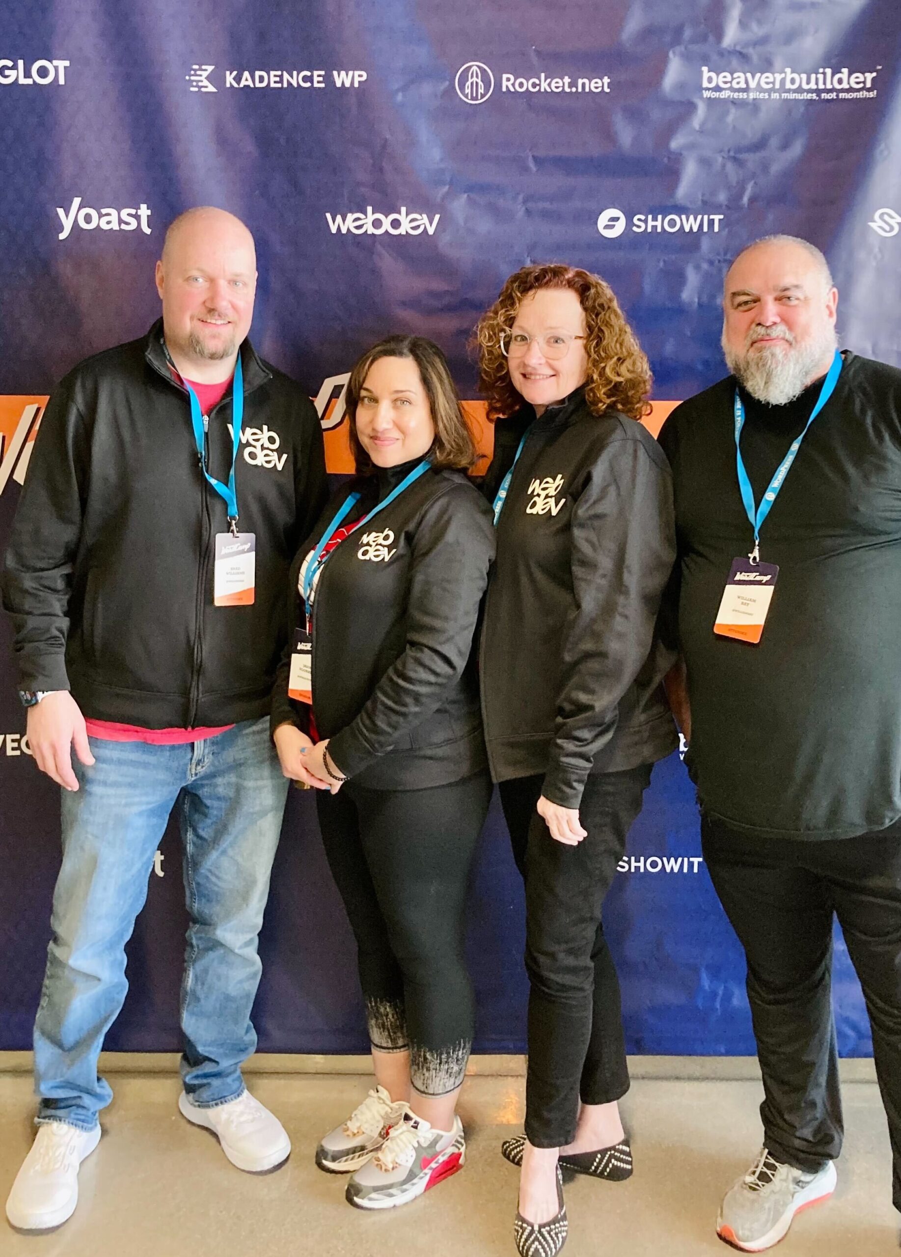 This is a photo of the team at WordCamp Phoenix standing in front of the event's backdrop, which features the WebDevStudios logo, among others. Standing left to right are CEO Brad Williams, Marketing Manager, Laura Coronado, Account Manager, Laura Byrne, and Frontend Engineer, William Bay.