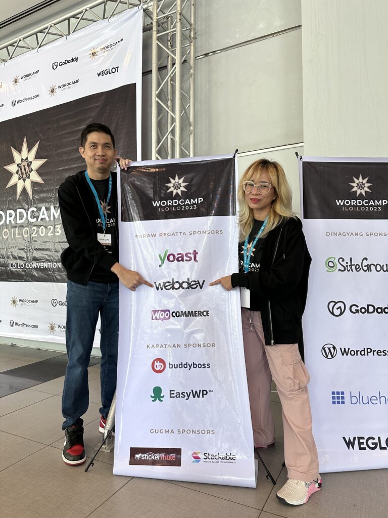 This is a photo of UX Designer Spencer Ravago and Engineering Manager JC Palmes standing next to the WordCamp Iloilo Sponsor Banner and pointing at the WebDevStudios logo.
