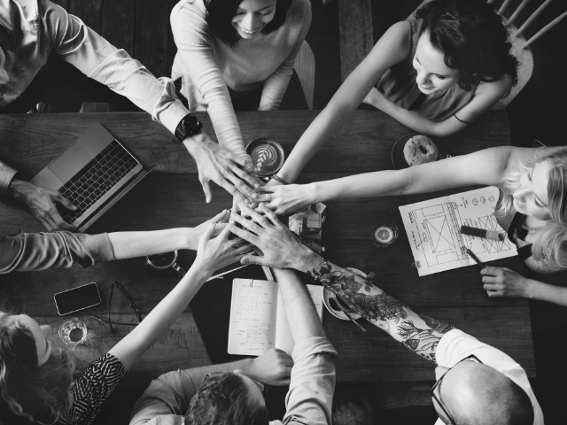 This is a black and white above view photo of a group of people at a table all reaching their arms and hands in to show unity.