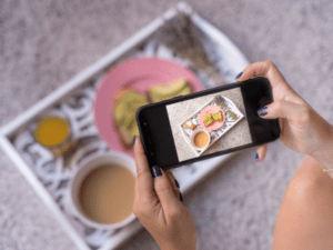 A pair of hands holding an iPhone to photograph a tray of food.